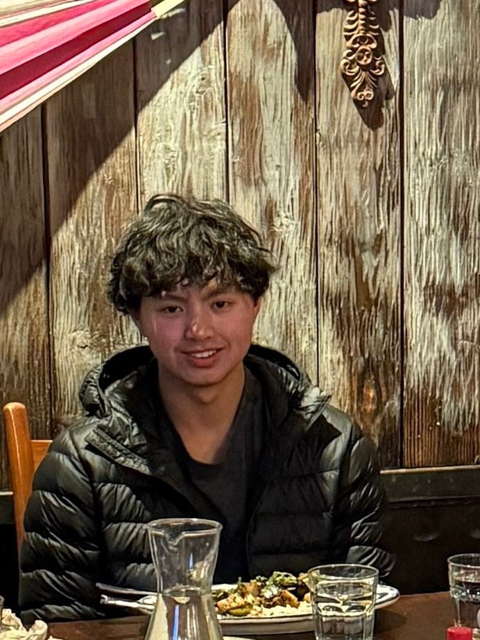 Person wearing a black jacket sitting at a wooden table with plates of food and a glass pitcher.